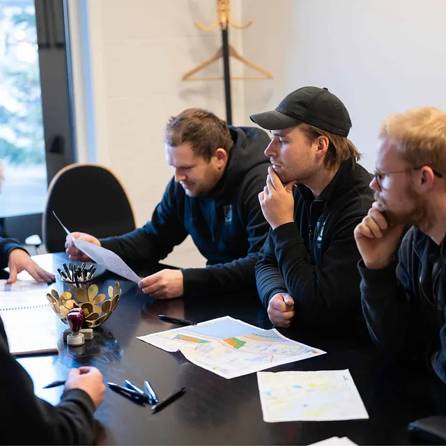 Fem menn i arbeidsmøte rundt et bord med byggetegninger og papirer.