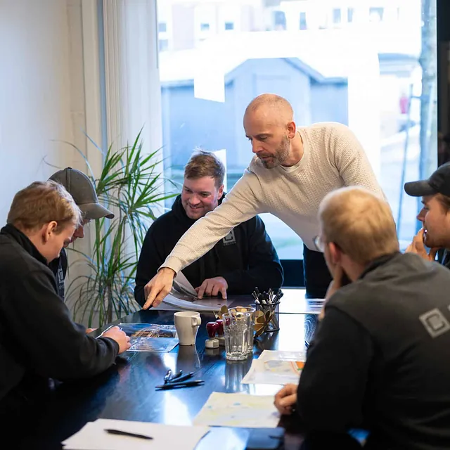 Mann peker på dokument under møte med kolleger rundt et bord med tegninger.