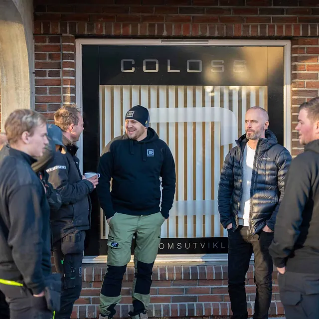 Gruppe arbeidere i samtale utenfor kontor med COLOSS-logo på vindu.