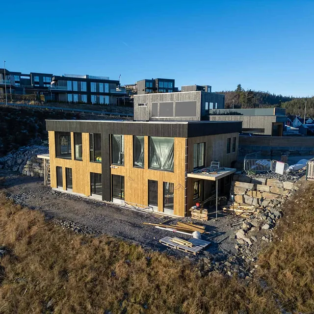 Moderne bolig med svart og naturfarget kledning i skrånende terreng.