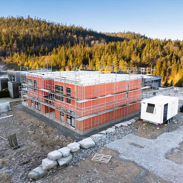 Oransje bygg under oppføring med stillas, brakker og skog i bakgrunnen.