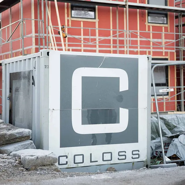 Container med stor COLOSS-logo foran bygg med stillas og rød isolasjon.